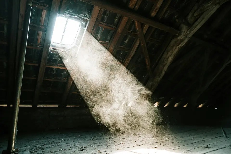 Een zolder onder een houten kap, met een bundel daglicht door het dakraam waarin stof door de lucht zweeft